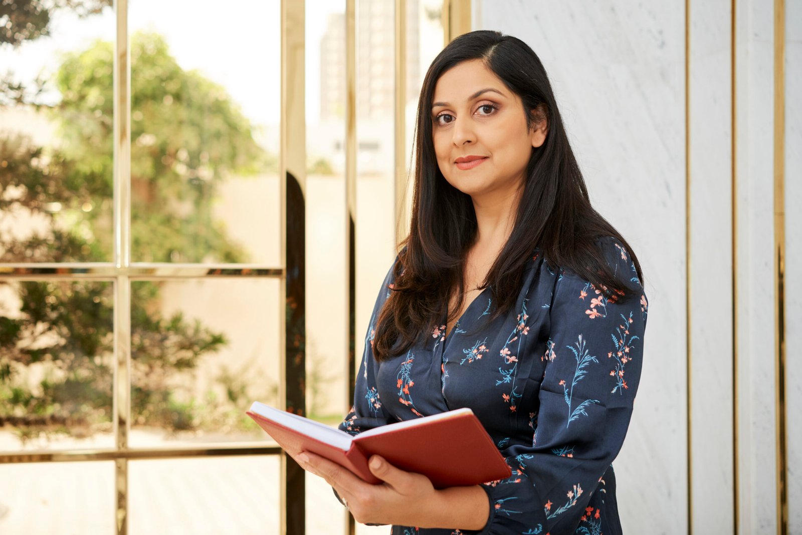Portrait of mature confident businesswoman standing near the window with notepad and looking at camera