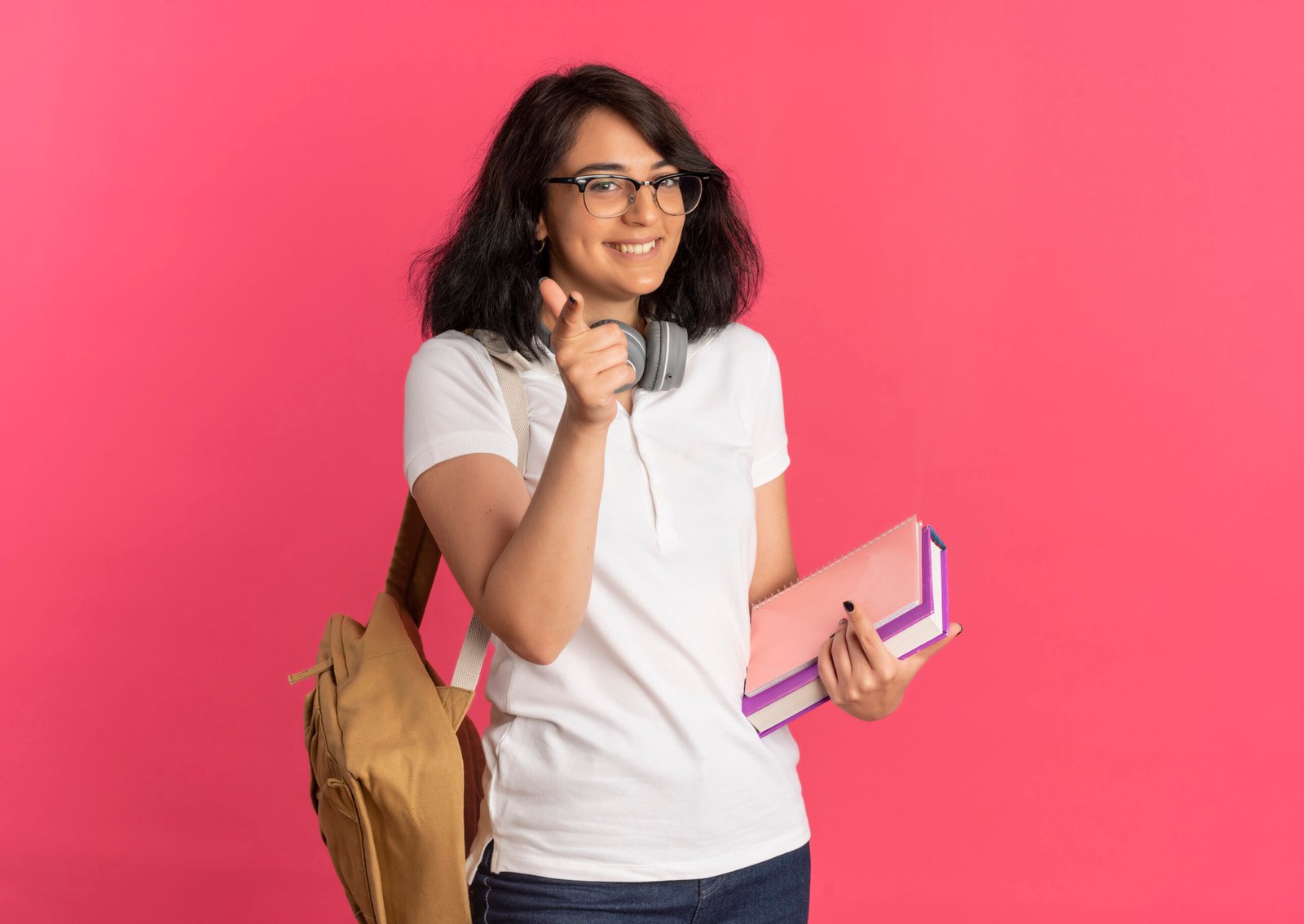 young smiling pretty caucasian schoolgirl wearing glasses and back bag with headphones on neck points at camera holding books isolated on pink background with copy space